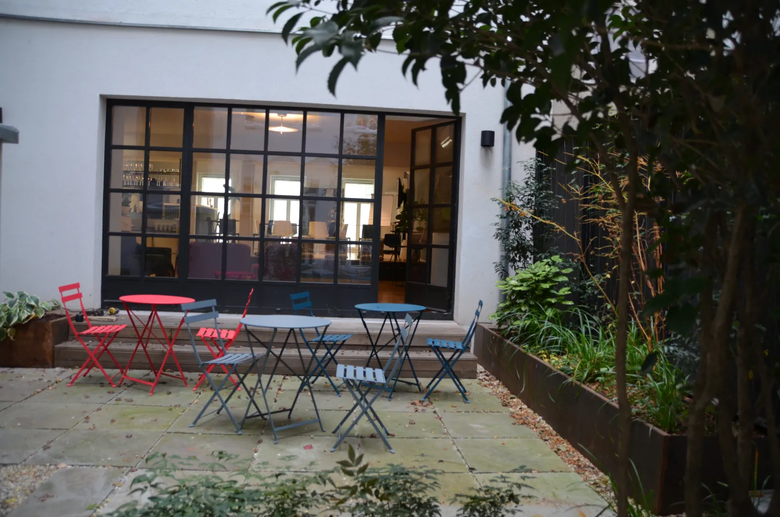 Blick vom Garten auf den Innenhof mit Bistromöbeln, Bäumen und Pflanzen